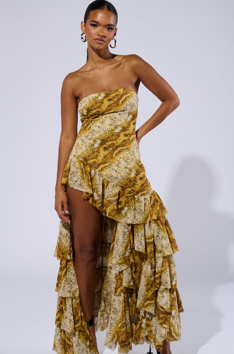 A woman models the TATTIANA SNAKE PRINT RUFFLED MAXI DRESS, which features a strapless design with multiple ruffles and an asymmetrical hemline. The dress showcases a striking yellow and white snake print pattern. She accessorizes with large hoop earrings and has her hair pulled back, standing against a plain, light gray background.