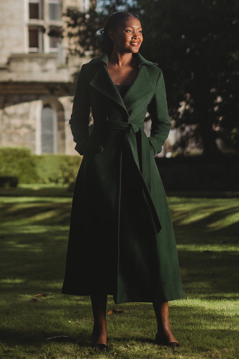A woman standing in an outdoor setting wearing a long green wool coat with double breasted design, a belt, and two side pockets. The coat has a mid calf length and long sleeves.