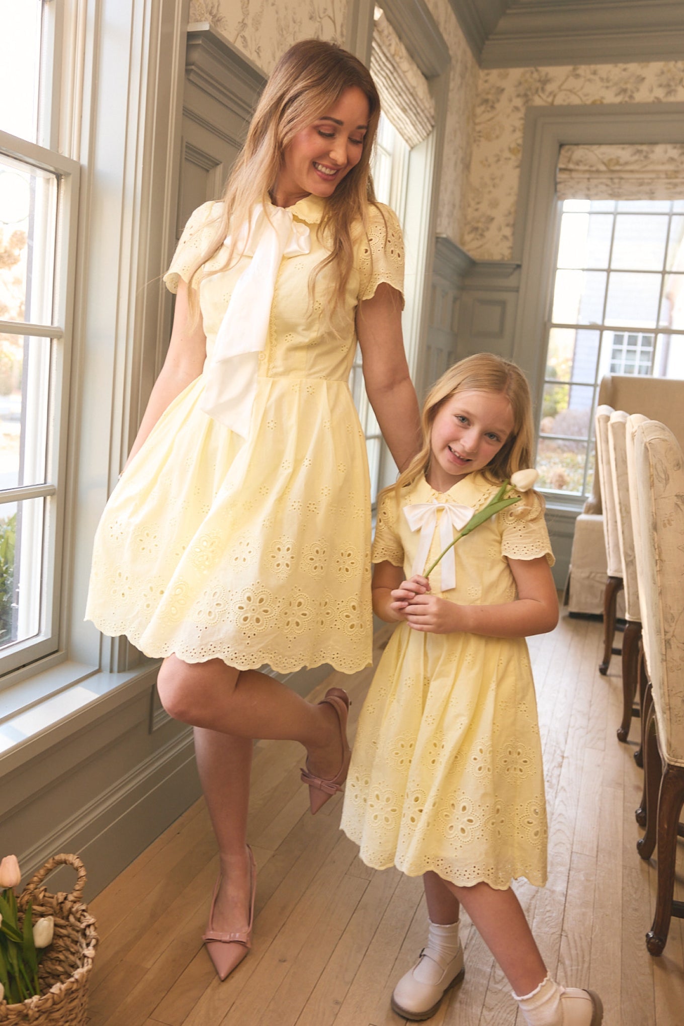 A woman and a young girl, both wearing Ivy City Co's Penny Dress in Butter Yellow Eyelet with white neck scarves, smile together in a bright room. The girl's holding a white tulip—these cotton dresses are perfect for warm days.