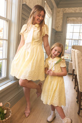 A woman and a young girl, both wearing Ivy City Co's Penny Dress in Butter Yellow Eyelet with white neck scarves, smile together in a bright room. The girl's holding a white tulip—these cotton dresses are perfect for warm days.