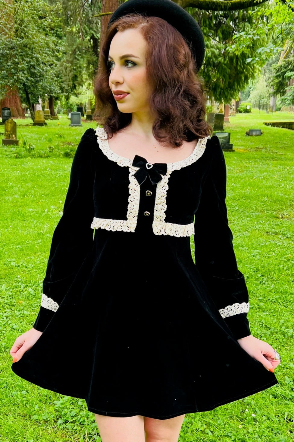 A woman in Witchwood Bags' Sabrina Luscious 60s style velvet mini dress stands on cemetery grass, looking to the side. She wears a black beret, has curly brown hair, and is surrounded by trees and headstones.