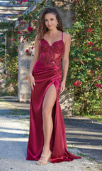 A woman stands outdoors by a green and pink flowered wall, wearing the Velvi Lace-Up Sheer-Corset Long Prom Dress Ivy—a sleeveless burgundy gown with a corset bodice and high slit.