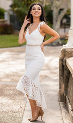 Woman outdoors in the Velvi High-Low White Graduation Dress Tahlia—white lace, tie straps, sleeveless high-low hem—posing and smiling with one hand near her hair; ideal for semi-formal events or as a chic white graduation dress.