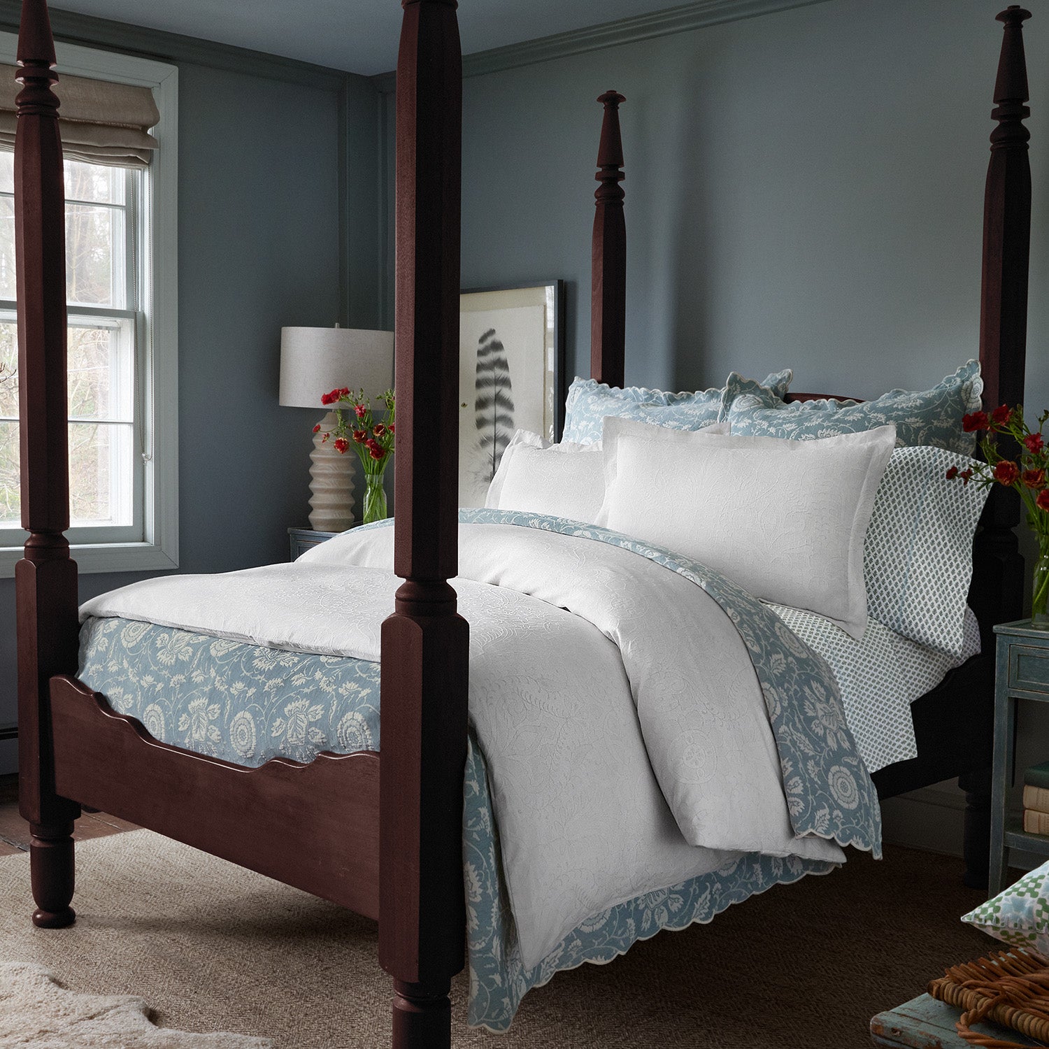 A bedroom featuring a dark wood four-poster bed adorned with the Kimari Mist Coverlet by John Robshaw in light blue and white jacquard cotton chenille, accompanied by a nightstand with a lamp, and vases of delicate woven florals on each side. The walls are blue-gray with a framed artwork near the bed.