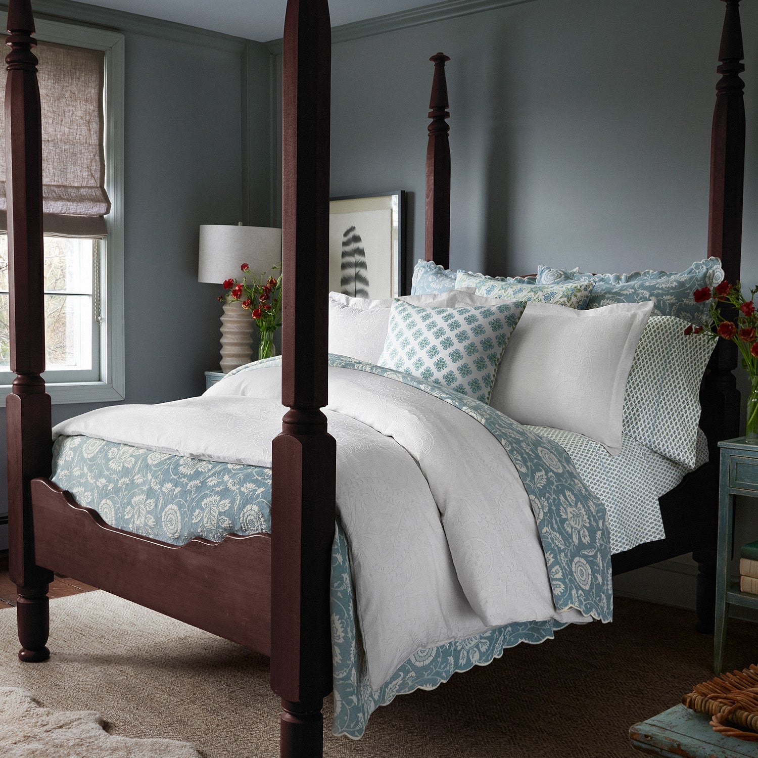 A neatly made four-poster bed with the John Robshaw Kimari Mist Coverlet, featuring delicate woven florals in blue and white, is centered in a room with light gray walls. A window with a beige shade and a small nightstand adorned with a lamp and flowers complete the serene setting.