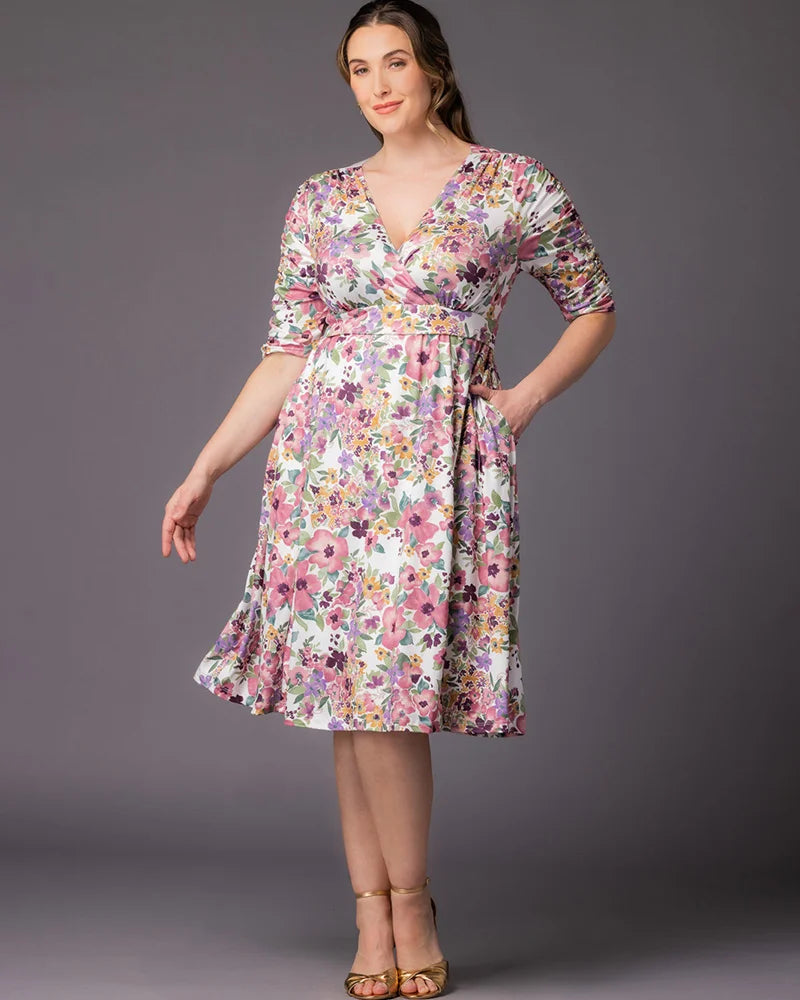 Gabriella Dress in Mauve Watercolor Bouquet Plus size model poses at a three-quarter angle against a soft gray studio background, one hand resting in a side pocket. She wears a white watercolor floral wrap-style midi dress featuring a deep V-neckline, ruched elbow-length sleeves, elasticized empire waist, and a full A-line skirt. Allover watercolor floral print features pink, lavender, purple, gold, and green blooms on a white background. Styled with gold strappy heeled sandals. Front view.
