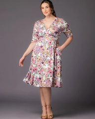 Gabriella Dress in Mauve Watercolor Bouquet Plus size model poses at a three-quarter angle against a soft gray studio background, one hand resting in a side pocket. She wears a white watercolor floral wrap-style midi dress featuring a deep V-neckline, ruched elbow-length sleeves, elasticized empire waist, and a full A-line skirt. Allover watercolor floral print features pink, lavender, purple, gold, and green blooms on a white background. Styled with gold strappy heeled sandals. Front view.