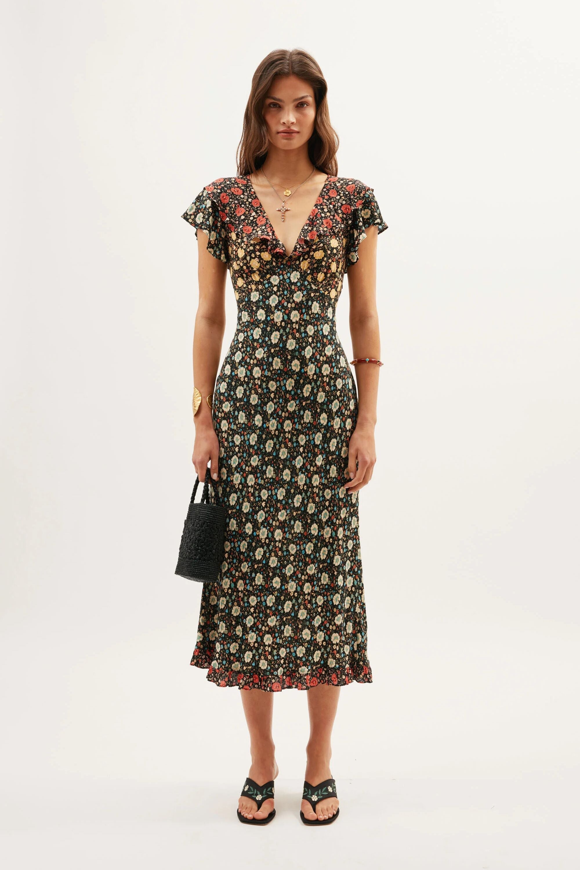 A woman faces forward in the Bethea - Eva Floral Mix Black, a fitted ankle-length crepe dress with a deep V-neck and flutter sleeves. She accessorizes with sandals, a black handbag, bracelets, and layered necklaces against a plain white background.
