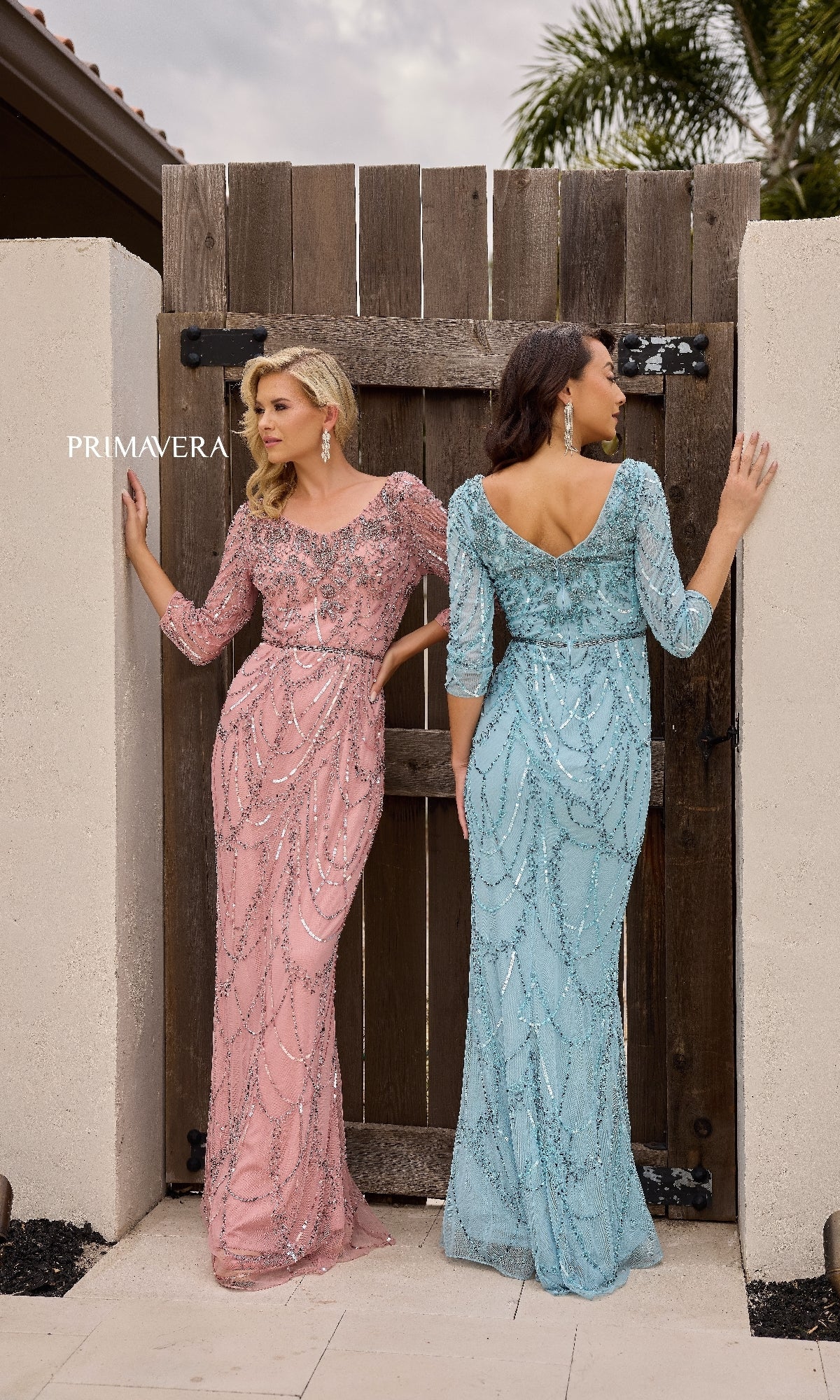 Two women stand by a wooden gate wearing Primavera 13156 long formal dresses—one in pink, facing forward, and one in light blue with a beaded waistline, facing away.