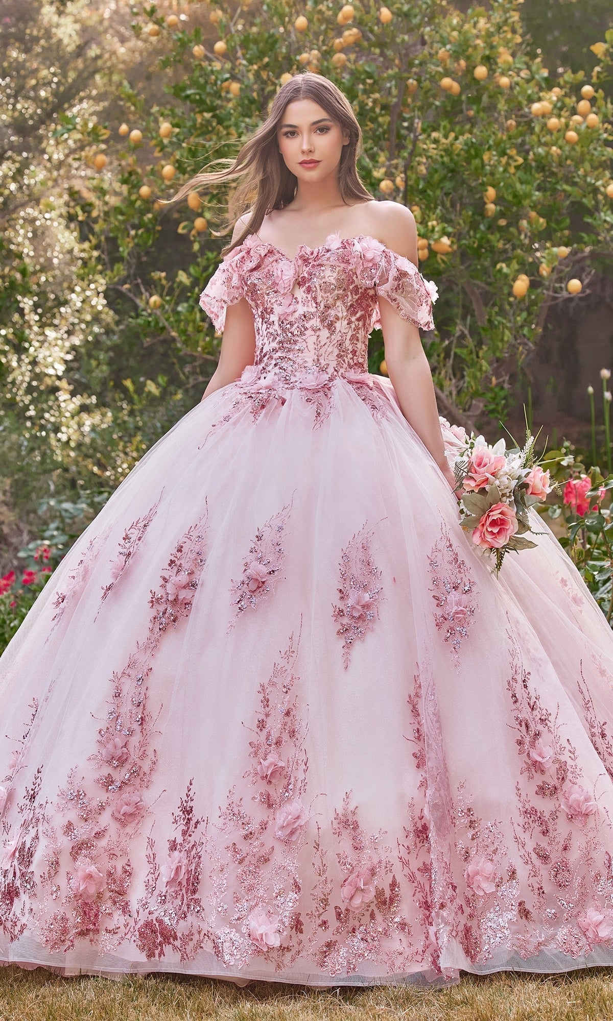 A woman stands outdoors in a garden wearing the Glitter Quince Dress with Back Bow: Ladivine 15701, a pink, off-the-shoulder ball gown with floral embroidery, holding a bouquet—an enchanting look perfect for a quinceañera.