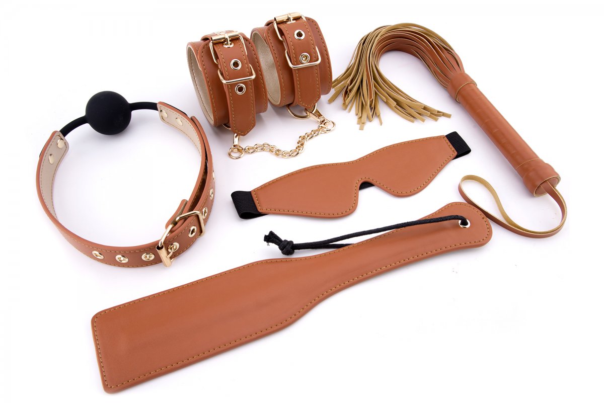 BDSM Set Veganskt Läder Cognac innehåller justerbara manschetter, en ögonbindel, ball gag, flogger och paddel, allt i brunt läder mot en vit bakgrund.