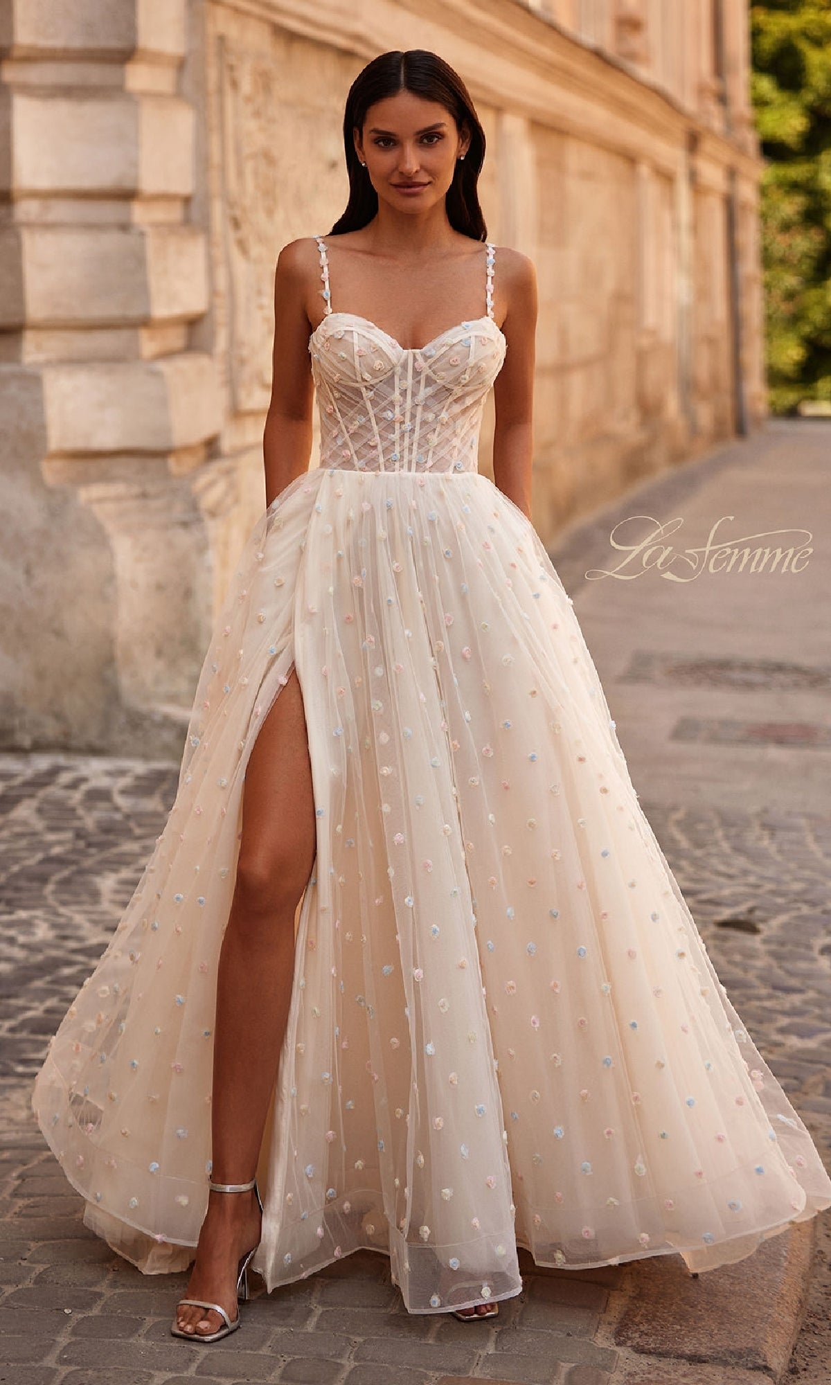 A woman in the cream Polka-Dot Long Prom Ball Gown La Femme 32716 with a sheer bodice, spaghetti straps, and high slit stands on a cobblestone street near a stone building.