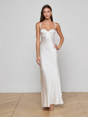A woman stands indoors wearing the Sayla Silk Gown, an elegant long white slip dress with thin straps and a fitted bodice. She poses with one hand on her hip against a plain white wall and wooden floor.