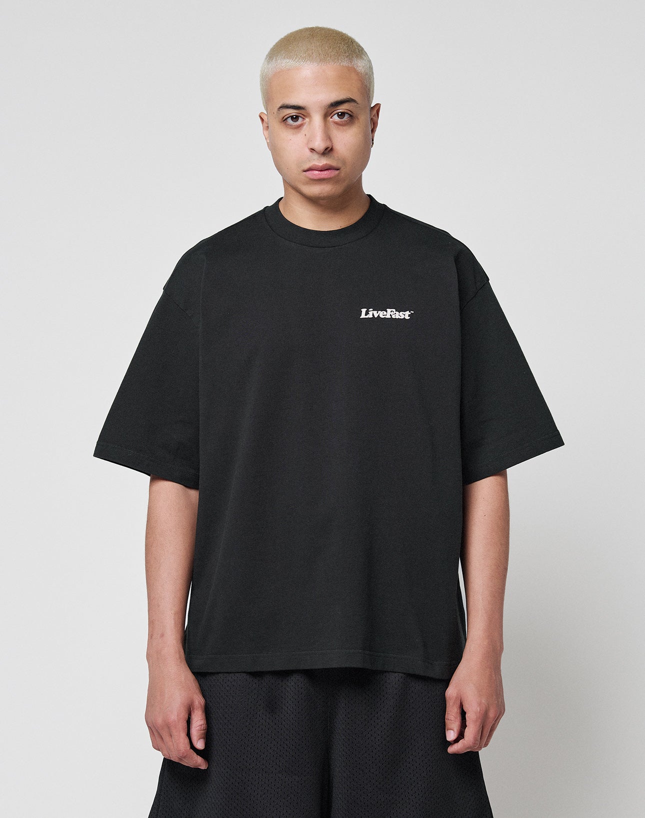 A person with short, bleached blonde hair wears the LIVE FAST DIE YOUNG Basic 280 Tee in black, featuring a small white LiveFast chest print and an oversize fit, paired with black shorts against a plain white background.