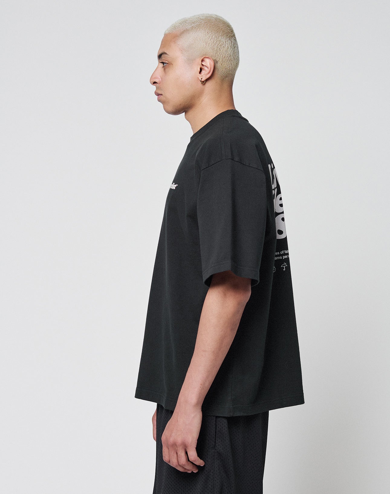 A person with short, bleached blonde hair stands in profile against a plain background, wearing the LIVE FAST DIE YOUNG Basic 280 Tee in black and matching shorts. The shirt showcases both front and back print details.