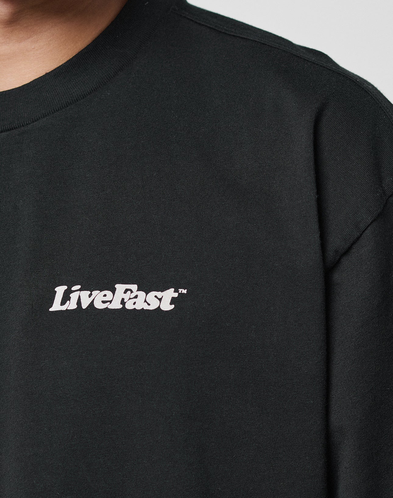 Close-up of a person wearing the LIVE FAST DIE YOUNG Basic 280 Tee in black, featuring white LiveFast™ text on the left chest. Only part of the face and upper torso are visible.