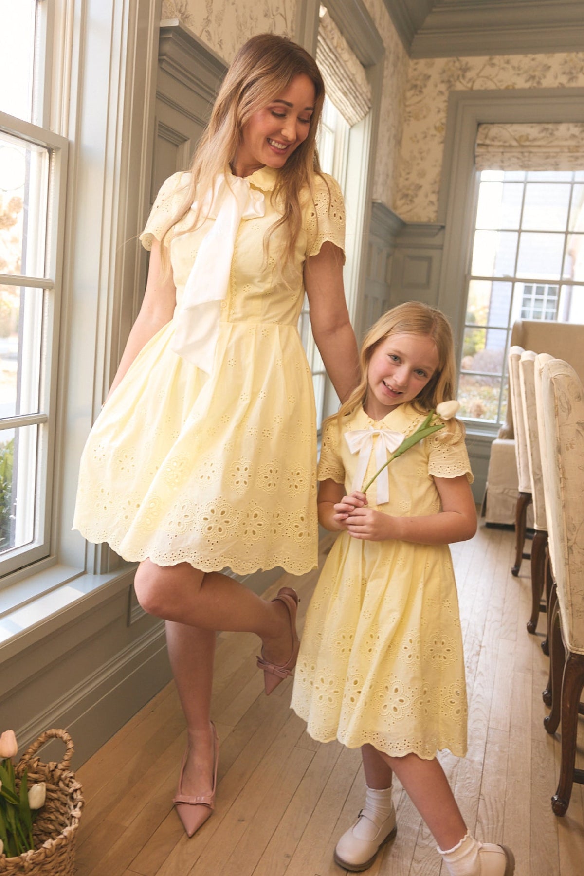 A woman and a young girl, both wearing Ivy City Co's Penny Dress in Butter Yellow Eyelet with white neck scarves, smile together in a bright room. The girl's holding a white tulip—these cotton dresses are perfect for warm days.