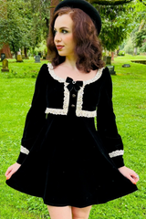 A woman in Witchwood Bags' Sabrina Luscious 60s style velvet mini dress stands on cemetery grass, looking to the side. She wears a black beret, has curly brown hair, and is surrounded by trees and headstones.
