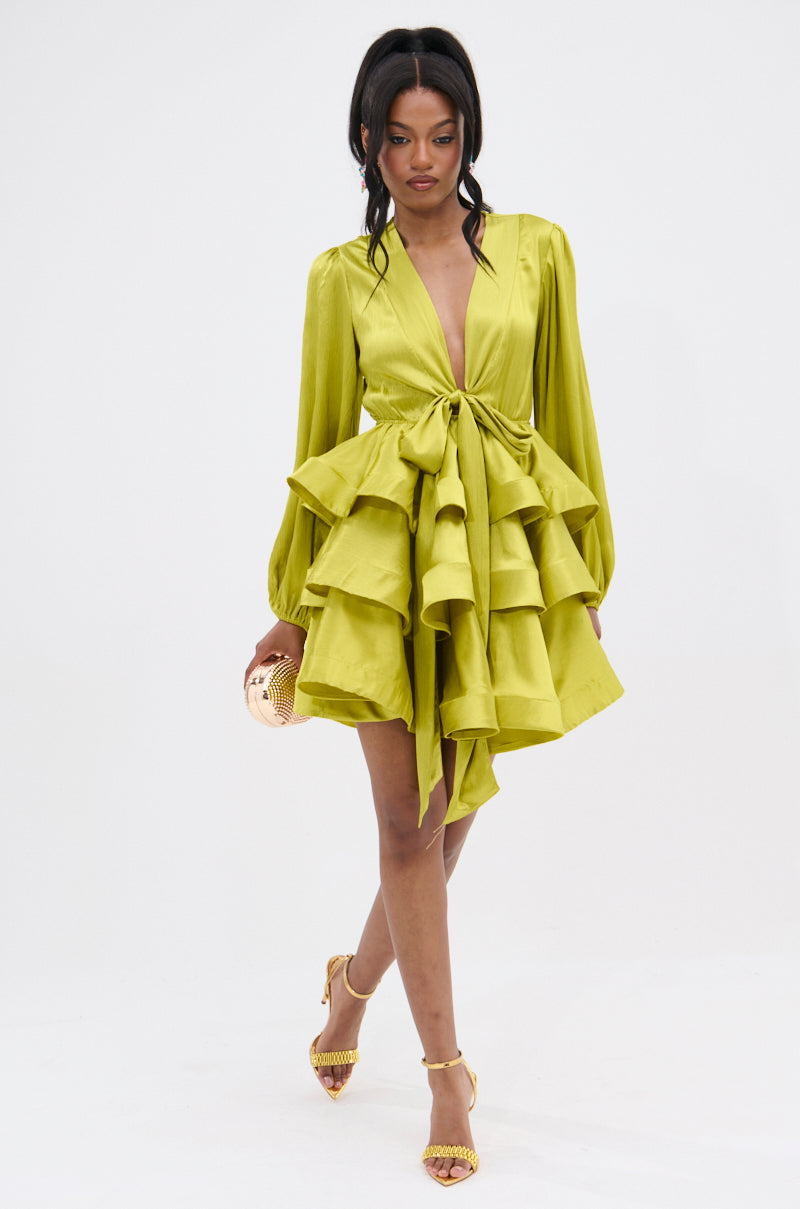 A woman wears the ARIANA SATIN RUFFLED MINI DRESS IN GREEN—featuring long sleeves, a deep V-neck, front bow, and ruffles—paired with a gold clutch and yellow heels, standing against a plain white background.