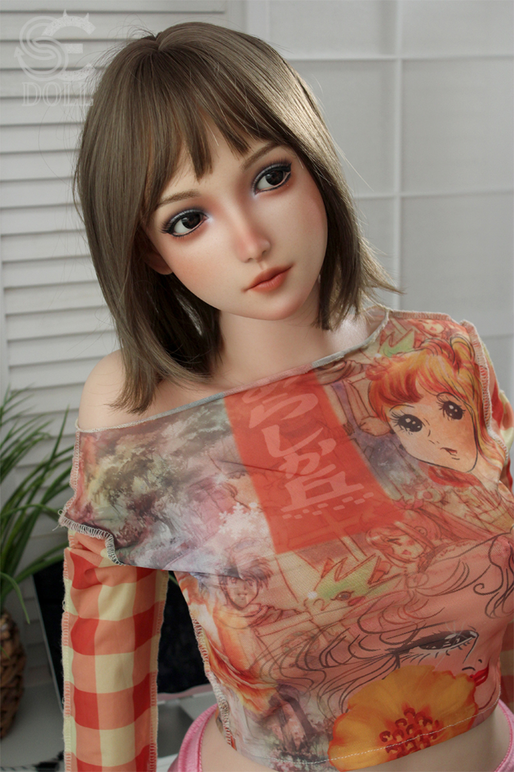 A tilted-angle portrait of the SEDOLL T160 cm B Silicone Pro - Yuuki.B wearing a vibrant graphic mesh top and pink bottoms against a neutral, white-paneled background.