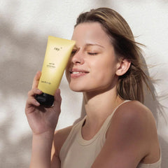 Een vrouw in een mouwloos topje lacht met gesloten ogen en houdt een gele tube Ray Zonnecrème SPF50 van Ray bij haar gezicht, tegen een lichte achtergrond.